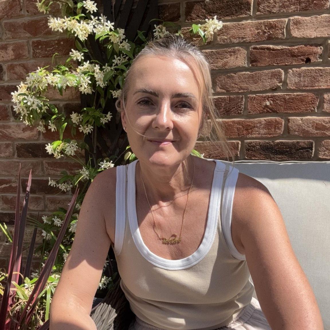 Natalie Heptinstall seated on a cushioned outdoor chair in the foreground, wearing a sleeveless light-coloured top and loose trousers. A necklace and bracelets are visible. Decorative plants, including a red spiky-leaved plant and a climbing plant with white flowers, surround the seating area. A brick wall forms the background.