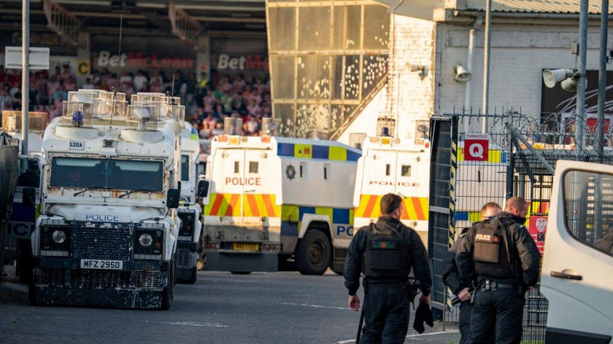Three police officers are standing next to a vehicle with a door open. A crowded stadium is behind them. Several police land rovers are parked.