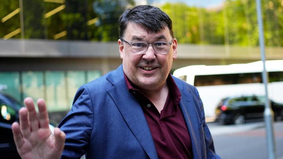 Linehan smiles and waves at the camera while standing outside in front of a road