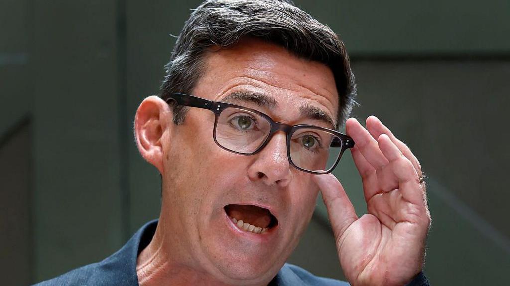 Andy Burnham making a speech. He is raising his hands up to his glasses. He has dark hair.