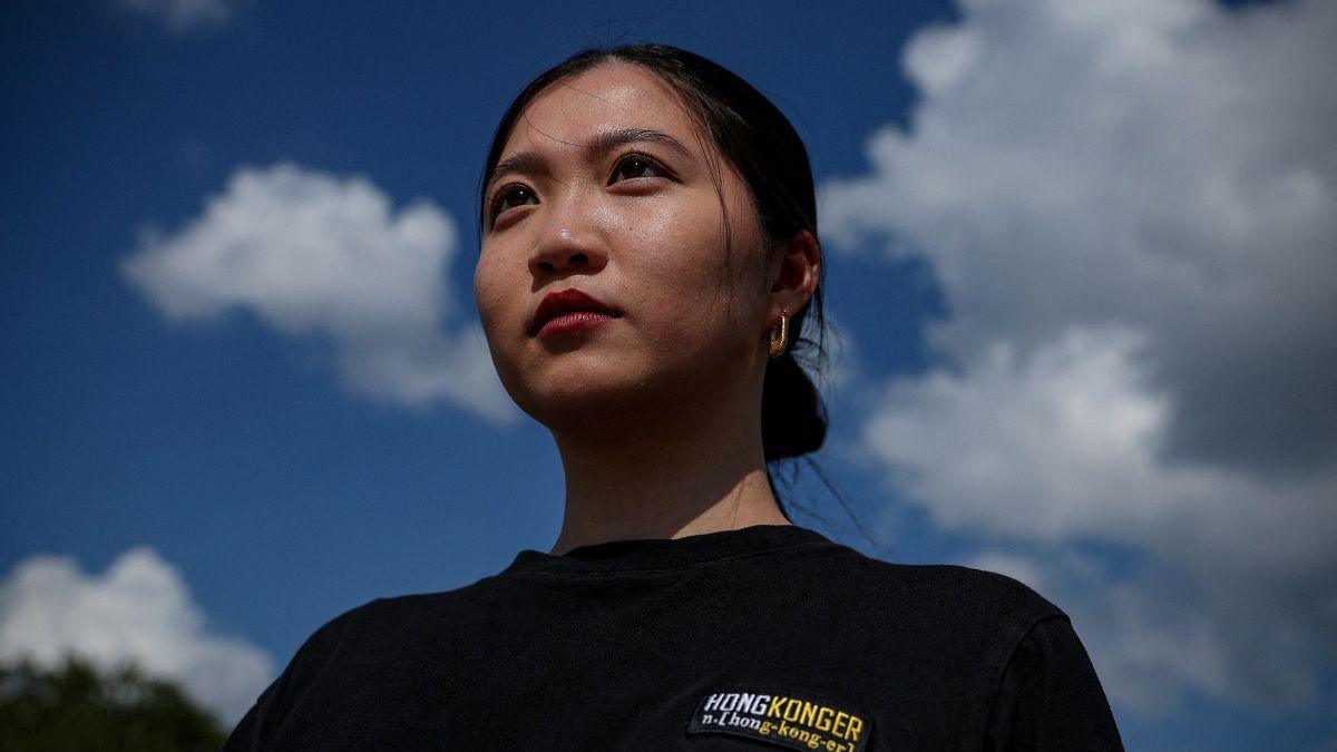 Anna Kwok looking ahead, wearing a black t-shirt. Her hair is tied into a bun. Behind her is a blue sky with clouds.