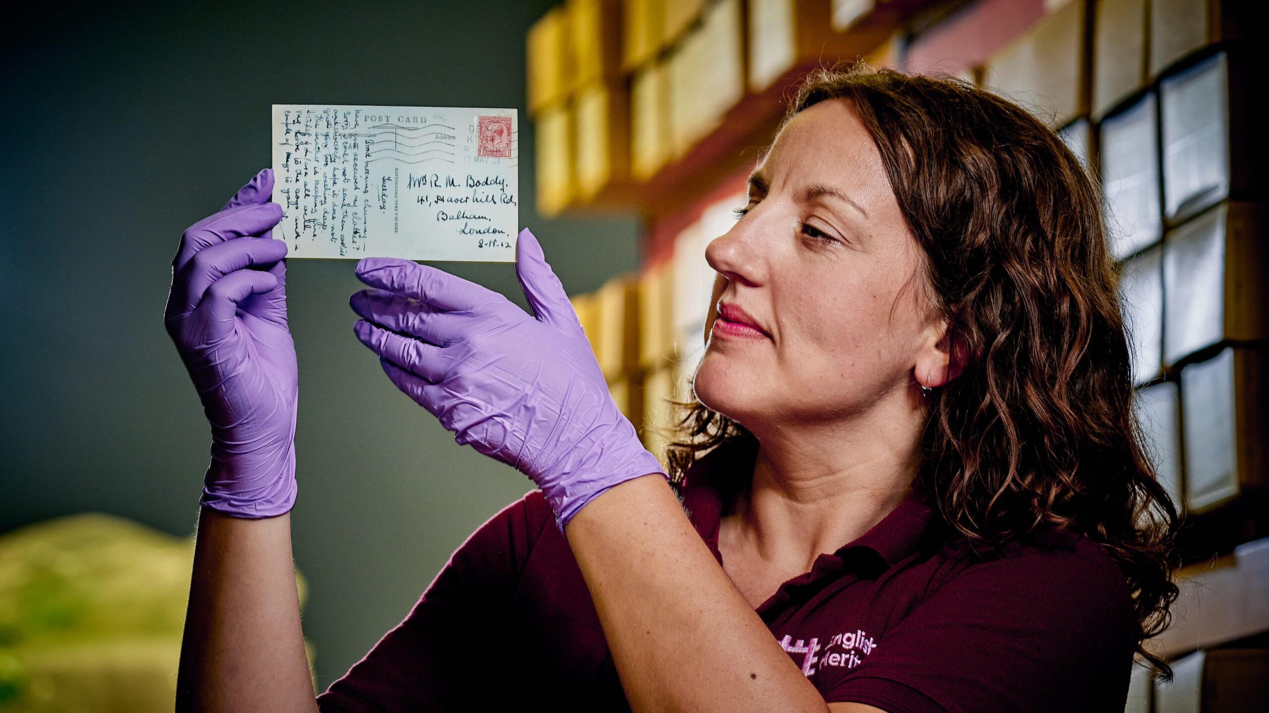 A woman wearing purple gloves holding a postcard. The handwritten back of the postcard is visible, which has a very old stamp and illegible handwriting.