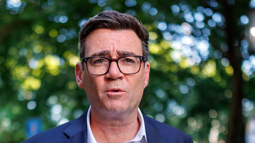 Andy Burnham in a white shirt and suit jacket. He is wearing glasses.