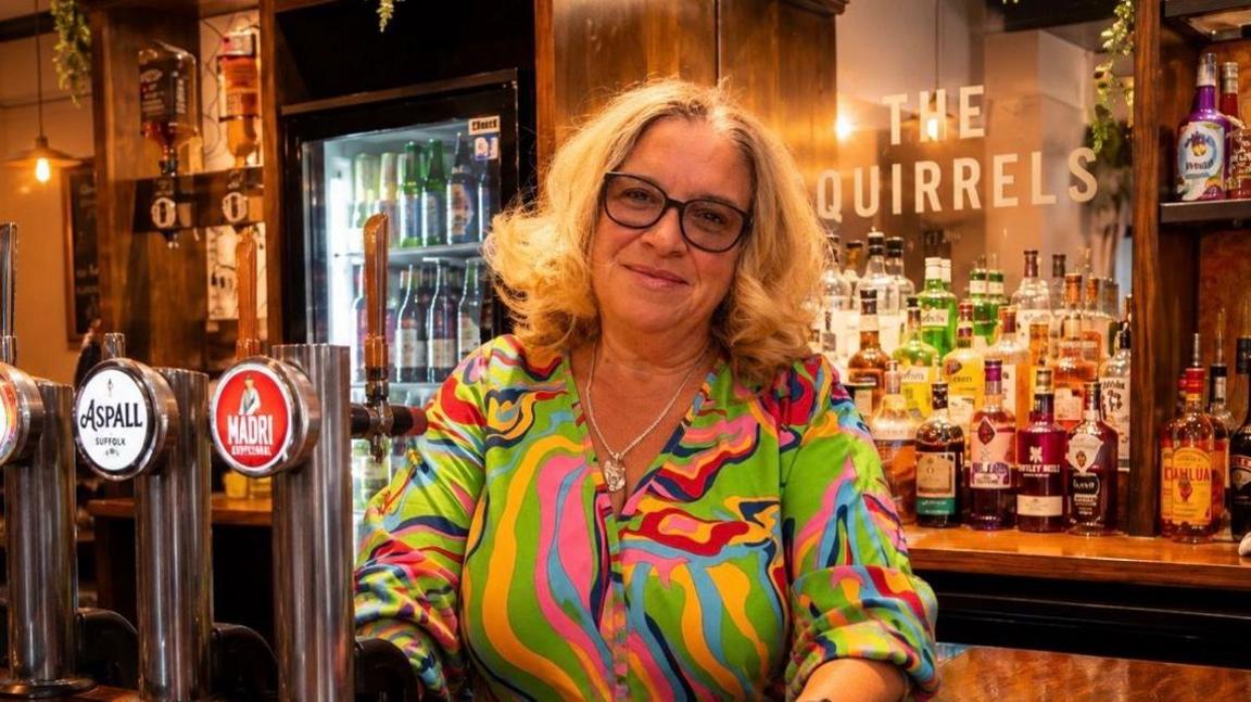 Miranda Richardson wearing a multi coloured shirt smiling at the camera as she stands behind her pub bar next to an Aspall and Madri beer tap with lots of spirit bottles behind her on a counter