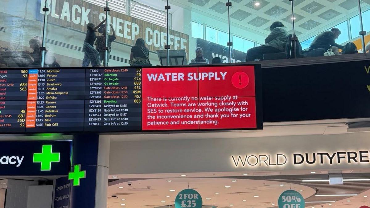 A red sign alerts passengers to the water supply issue on a screen in a terminal building. A World Duty Free sign is underneath and a pharmacy sign is nearby. A further screen shows flight information.