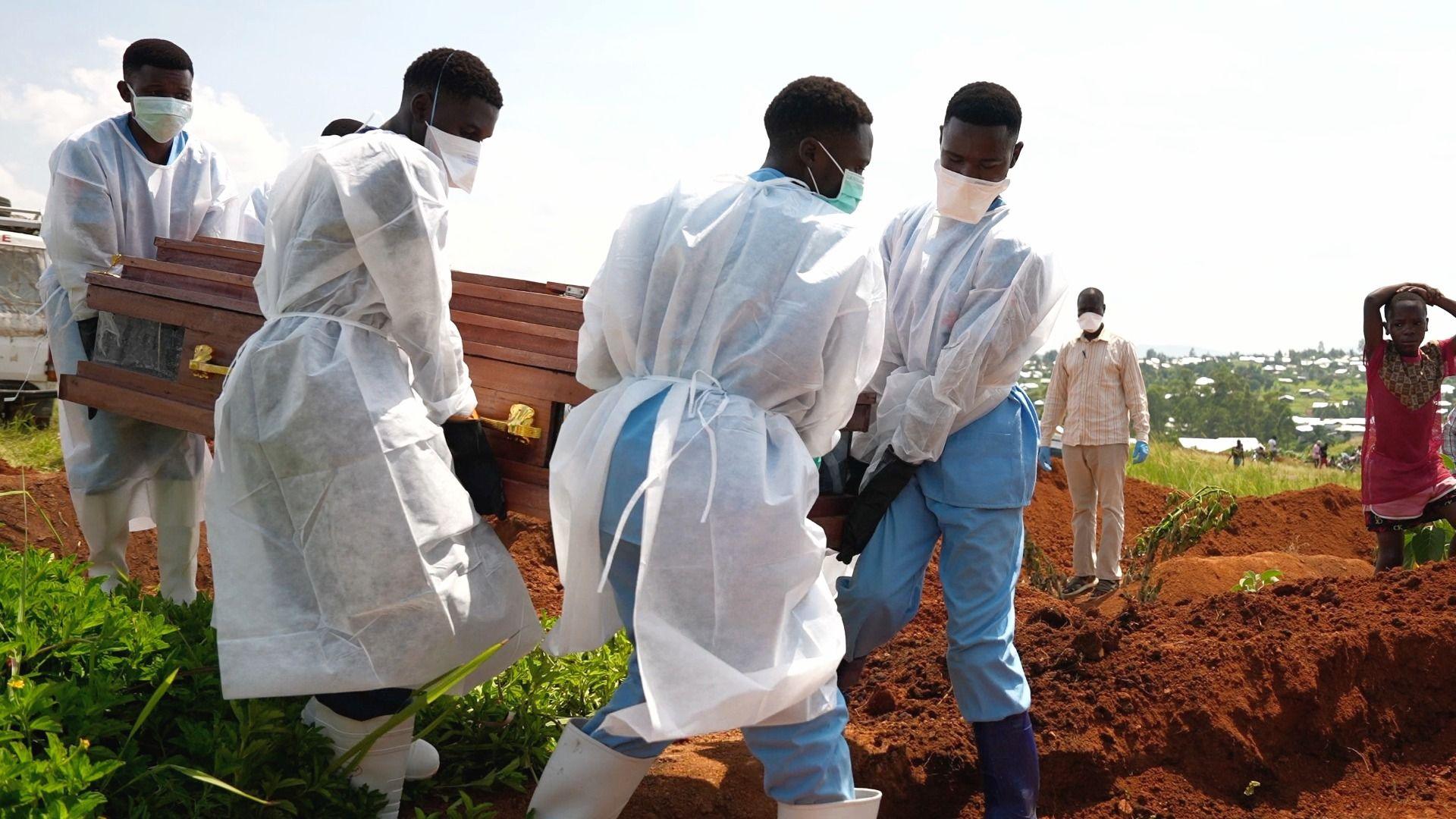 Volunteers in blue scrubs, rubber boots and white gowns carry a coffin towards a dug grave. A few people, including one holding a spade, watch on from the side.