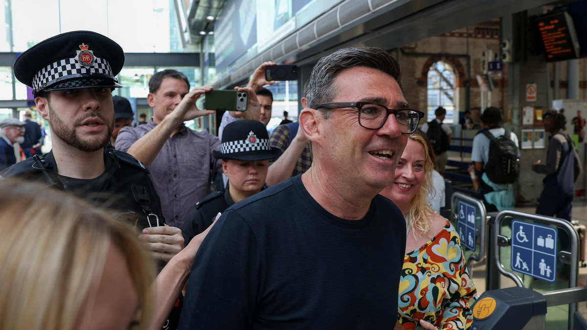 Andy Burnham flanked by two police officers. There are crowds of people taking photos behind him. He is wearing glasses and a dark shirt.