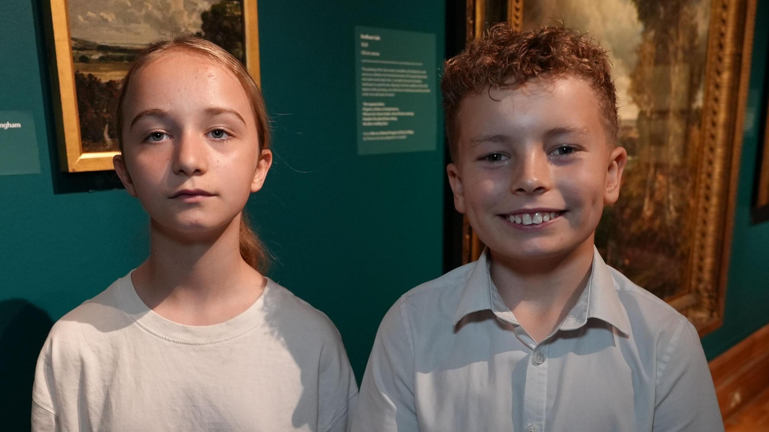 A young school girl and boy smile at the camera in an art exhibition. The girl has blonde hair that has been tied back and is wearing a white T-shirt. The boy has curly short red hair and is wearing a white school shirt.