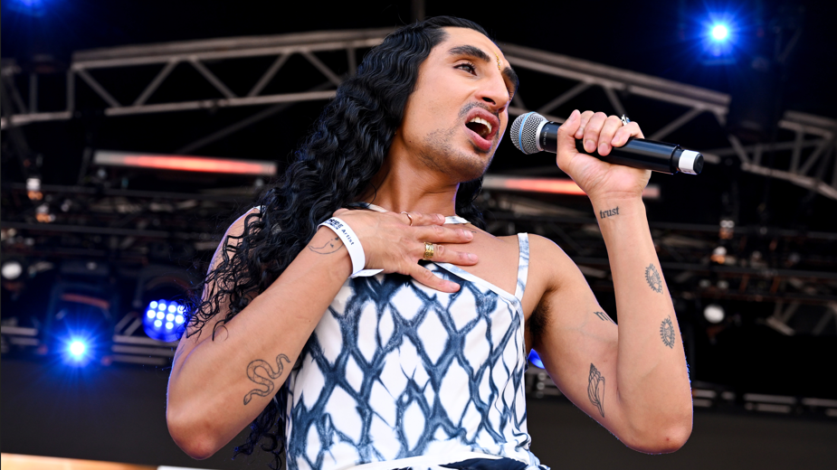 Shot of singer Leo Kalyan on stage at the Trafalgar Square Show on Saturday 4 July. He wears a white and black strappy top and sings into the mike. He has long dark hair and light beard growth.