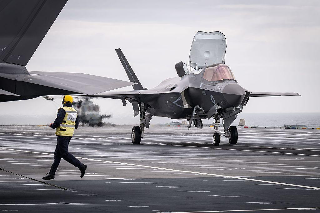 A member of the flight deck crew runs past as an F-35B, the STOVL (short takeoff and vertical landing) variant of the Lockheed Martin F-35, takes off from the flight deck of HMS Prince of Wales (R09), a Queen Elizabeth-class aircraft carrier