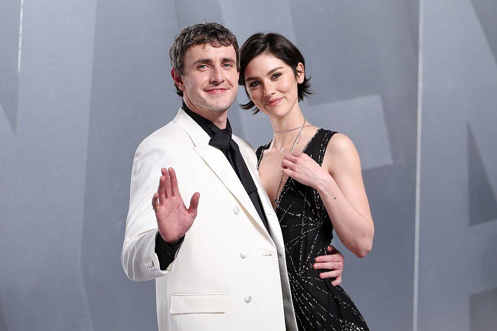 Gracie Abrams and Paul Mescal on the red carpet of the Vanity Fair Oscars Party in 2026. Mescal is wearing a white suit, and waving at the camera. Abrams smiles as the actor puts his other arm around her waist.