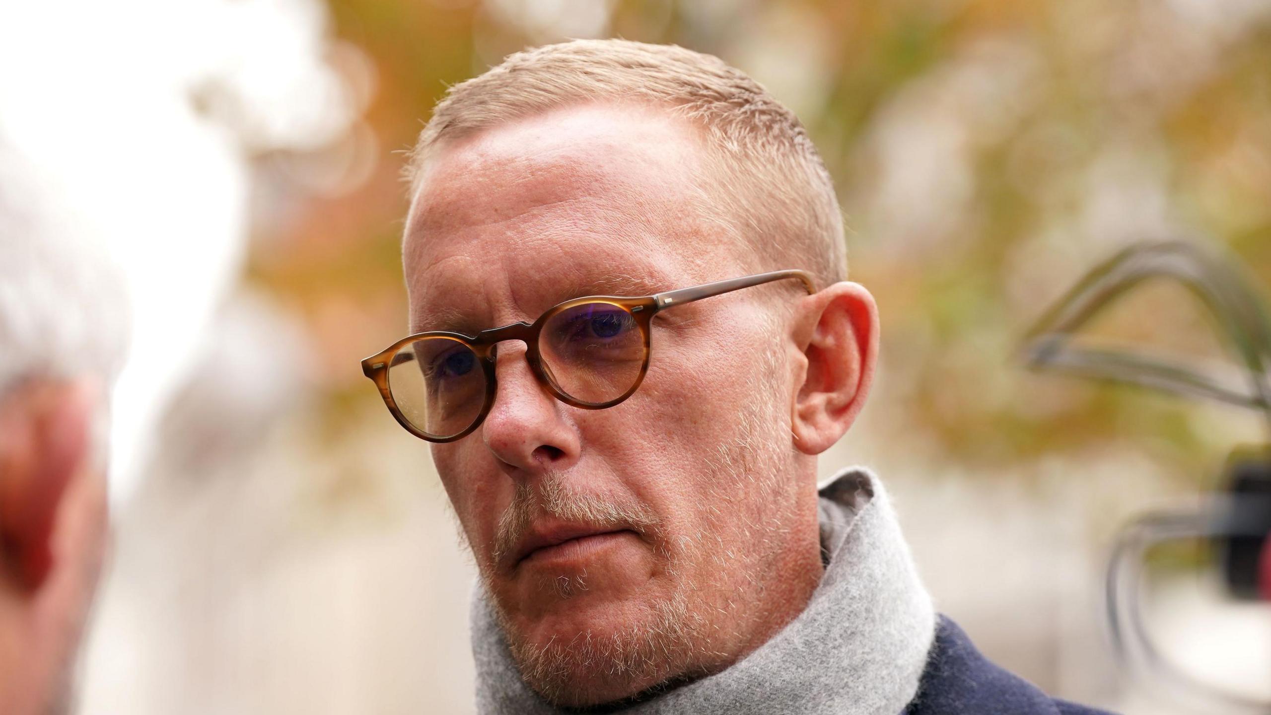 Laurence Fox has short blond hair and stubble. He is wearing round-framed brown glasses and a grey scarf over a navy blue coat.