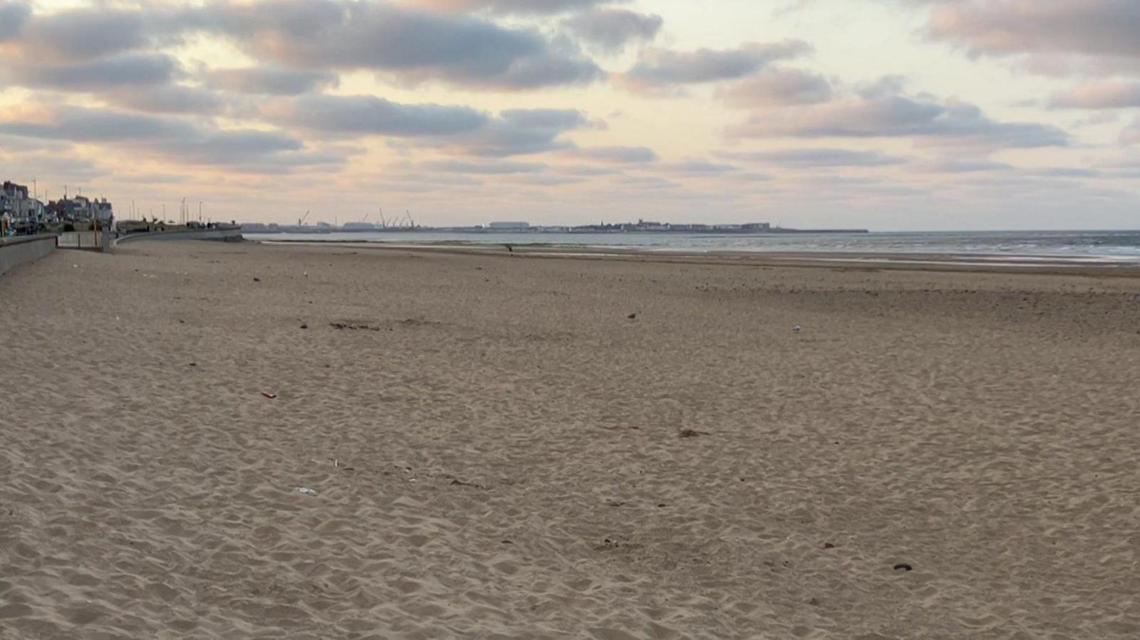Long shot of a beach as the sun is setting. The beach is sandy and you can see the sea in the background.