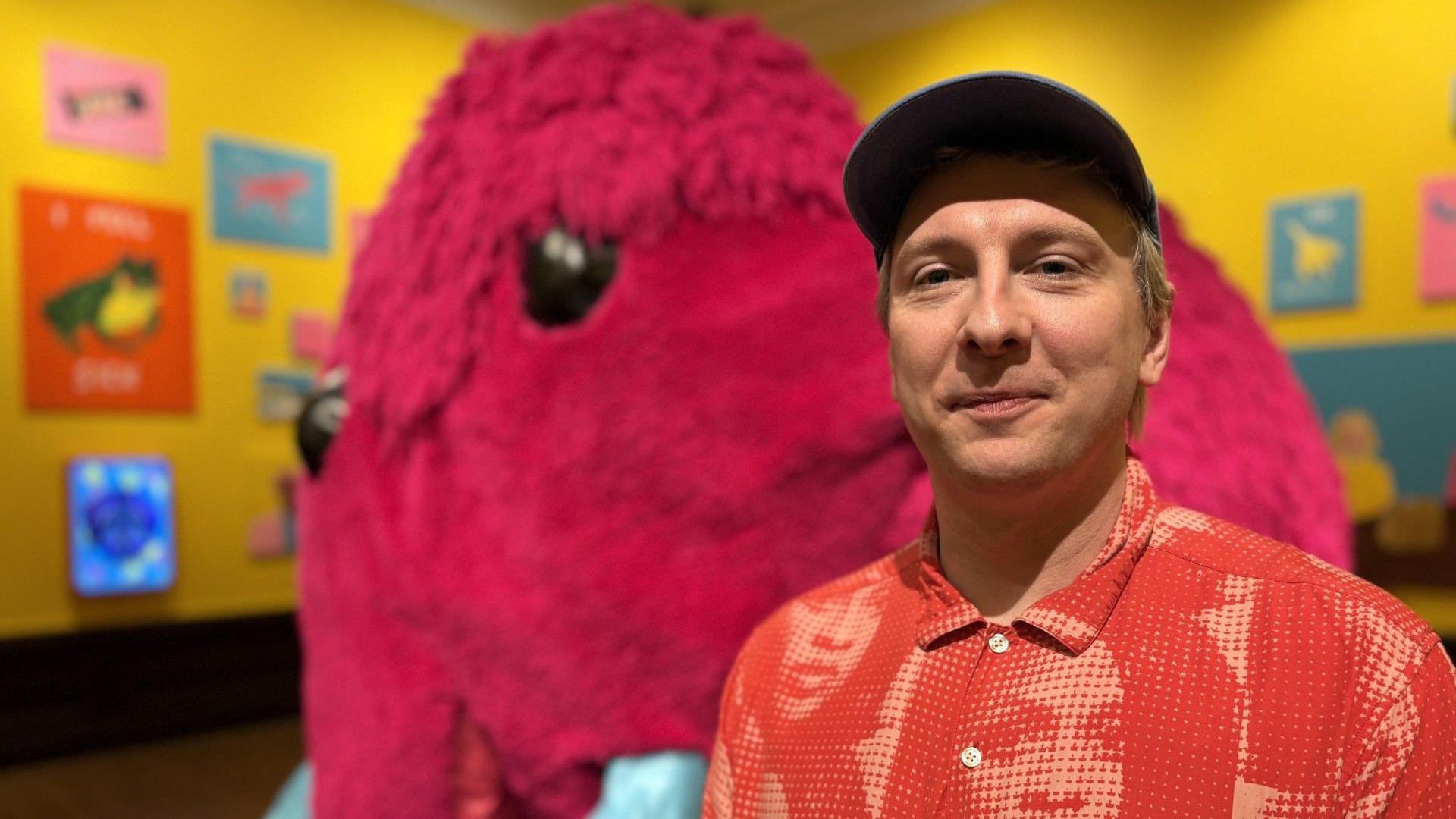 A man in a red shirt and cap smiles at the camera as he stands in a room with yellow walls, colourful paintings and a pink woolly mammoth sculpture behind him.