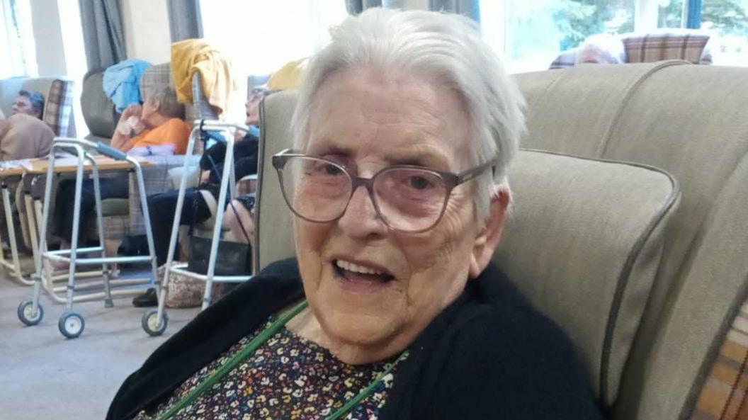 Lisa Greenwood's mother Mavis is pictured smiling at the camera in a care home setting. She is wearing glasses, and a dark cardigan over a dress.