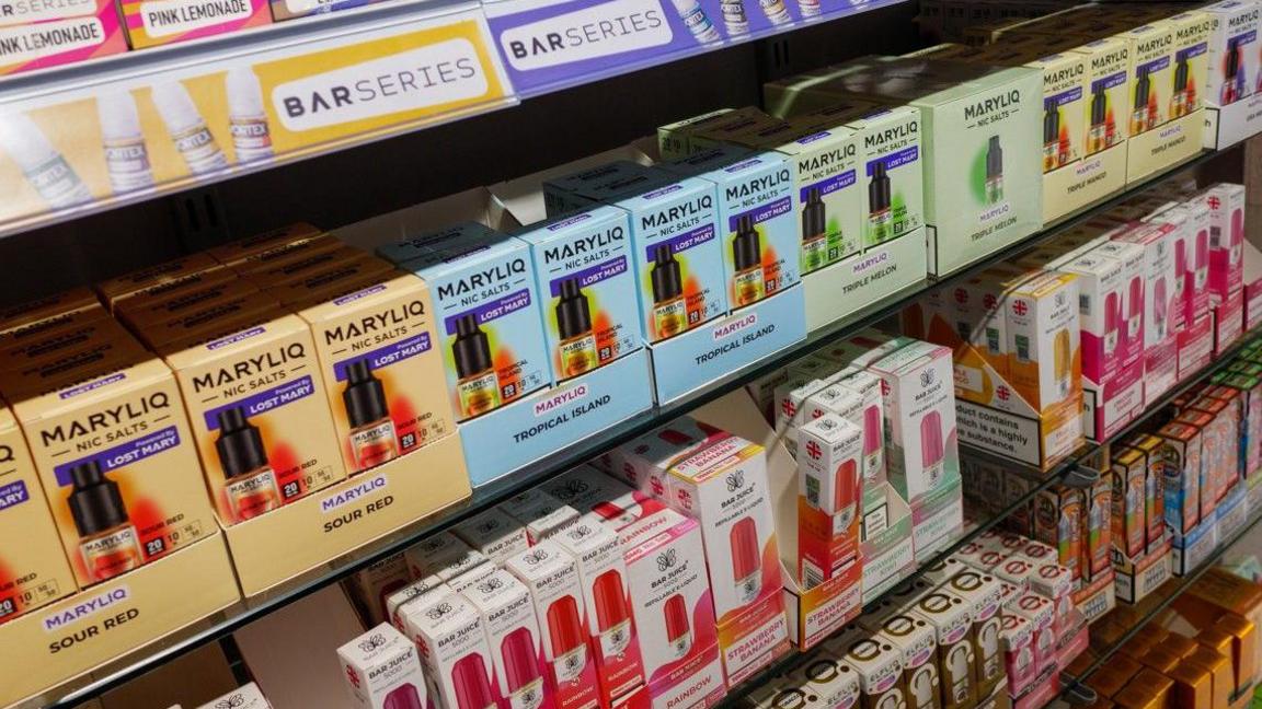 Several shelves, presumably from the inside of a shop, are full of boxes of refillable vape juice. Many of the boxes are in bright colours like pink, yellow and blue.