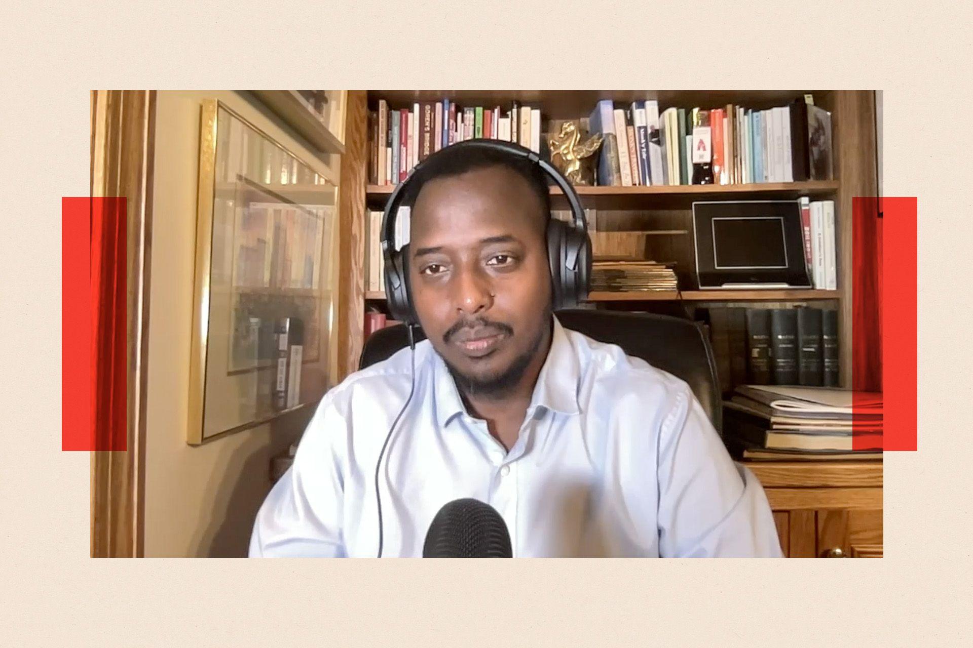 A man in an office wearing over-ear headphones and a white shirt, talking into a microphone