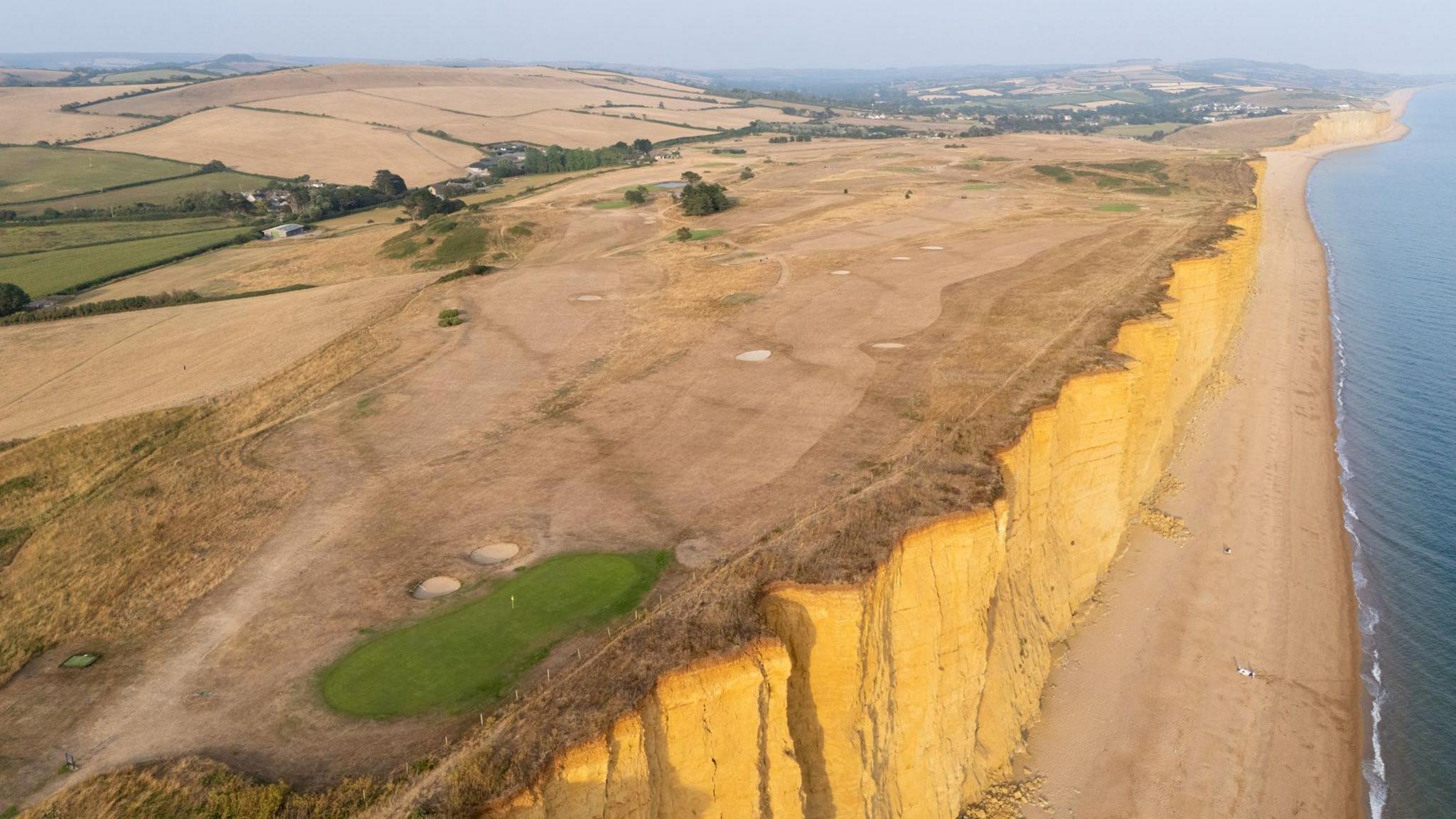 An aerial view of a clifftop and a golf course and fields to the left. The grass is brown due to drought and the sky is grey in colour.