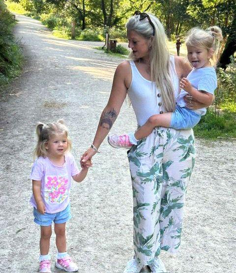 Brioney carries one twin while holding the other's hand while walking along a grey, stoney path bordered by greenery and trees in the sunshine - they all have blond hair