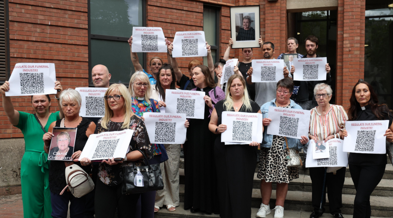 A group of women and men stand on steps outside a red-brick courthouse. Each of them holds a sheet of paper with a QR code and the words "Regulate our funeral industry" written on them. Some of them are also holdings photographs of loved ones