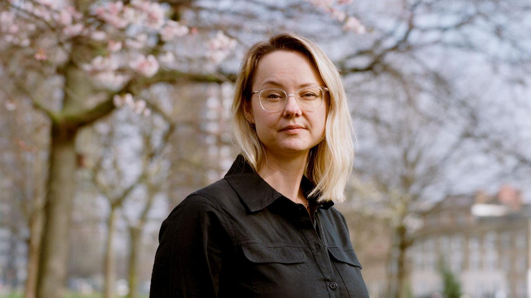 Cara Cinnamon is wearing glasses and a black button-up shirt, standing outdoors beneath blooming tree branches. The portrait is framed from the waist up, with a softly blurred background of trees and buildings in a park-like setting under natural daylight.
