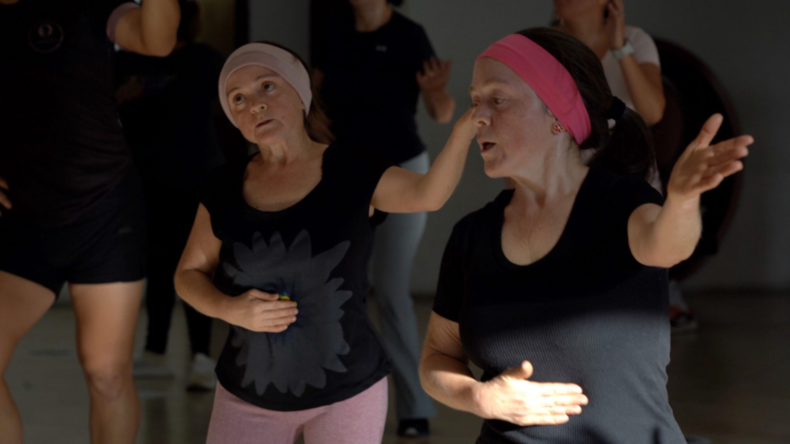 María Luisa and María del Cisne at a dance class - they both hold one hand across their stomachs with the other in the air. They both wear black t-shirts and pink hair bands