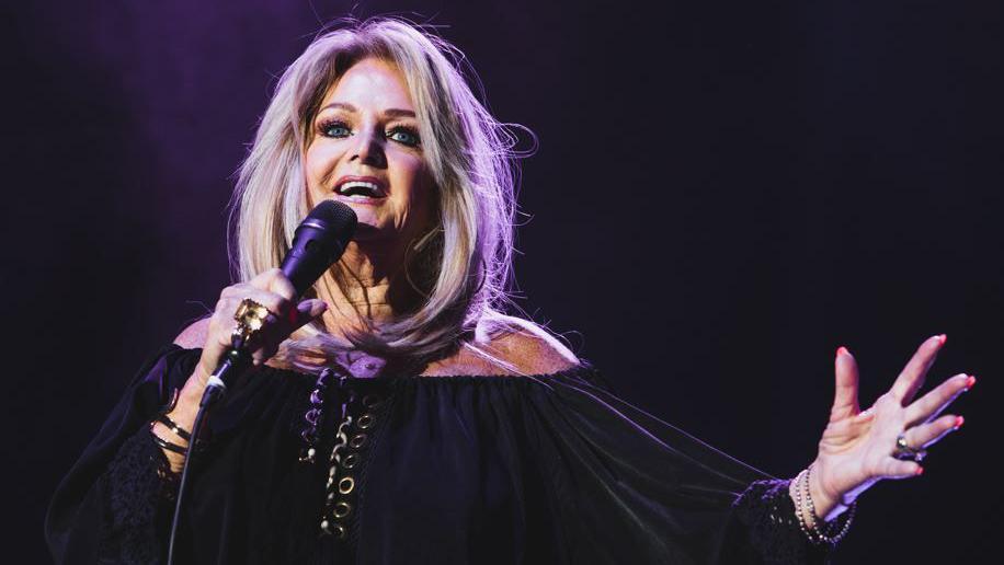 Bonnie Tyler on stage singing. She wears a black long sleeve dress and holds the microphone in one hand, with the other outstretched. Her long blonde hair is loose.