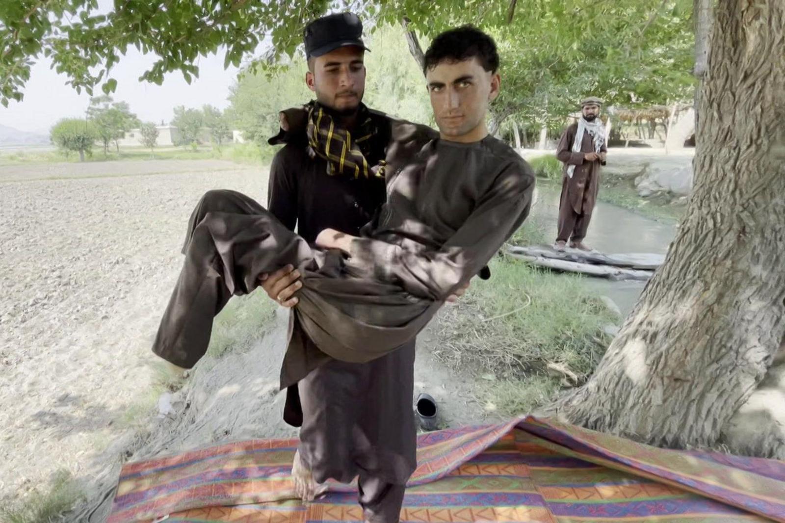 One of the 12 migrants who spoke to BBC Afghan Forensic, quadruple amputee Shahsawar, 21, is carried by another man, on a red-and-blue striped rug, while a third looks on, in a tree-lined Afghan landscape. All wear brown salwar-kameez, the two unnamed, bearded, men hats and patterned scarves.