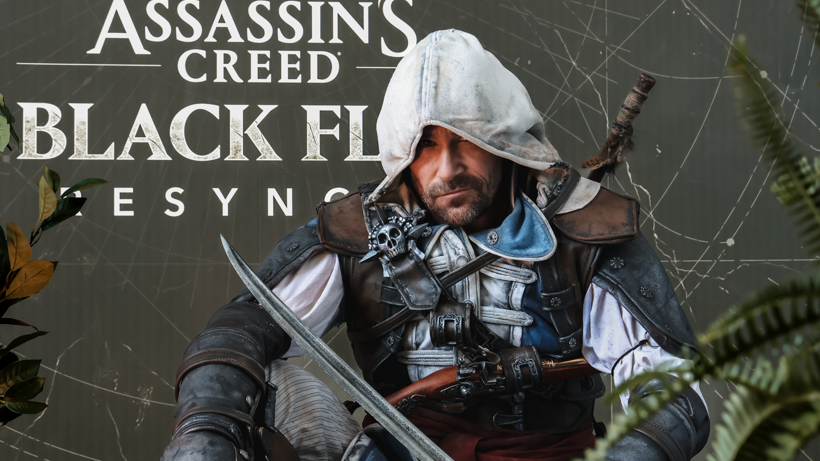 A man with a dark beard is sitting in pirate-style costume with his white hood up, holding a sword. He is learning against a black background looking at the camera. Another sword can be seen strapped to his back, with a pistol on a harness on his chest.