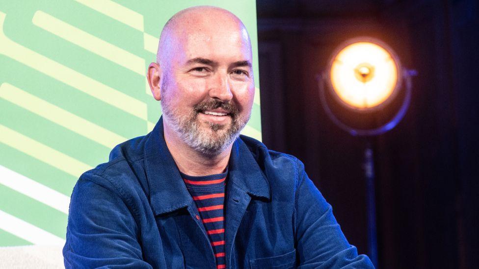 Scottish-American writer and fashion designer Douglas Stuart attends the Edinburgh International Book Festival at Edinburgh College of Art on August 28, 2022 in Edinburgh, Scotland. He is waring a blue and red stripey T-shirt and a blue jacket.
