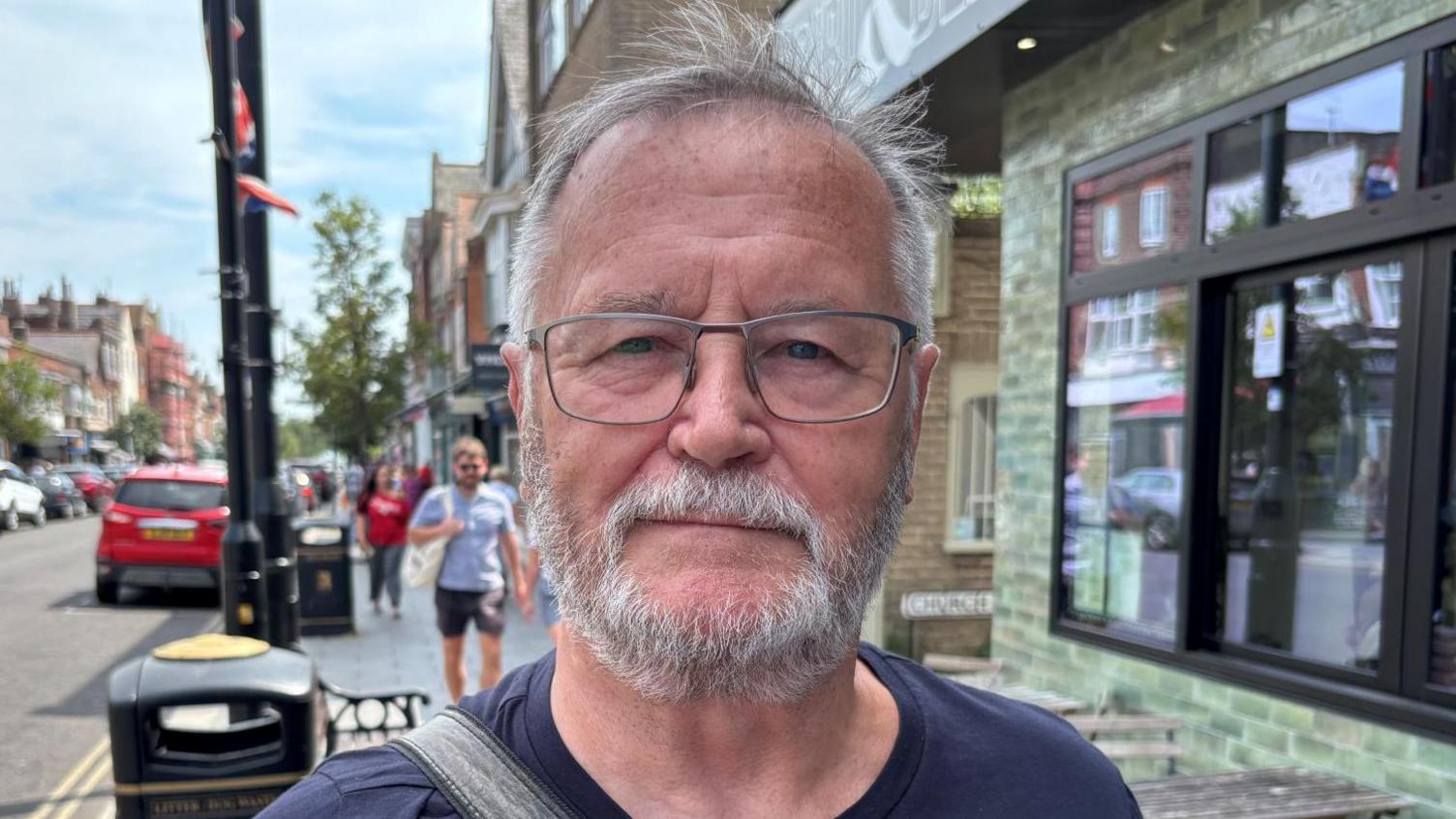 Graham Smith has short grey hair and a beard. He is wearing a navy T-shirt and glasses on Connaught Avenue, which is Frinton-on-Sea's high street.