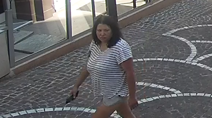 A woman with dark hair and a serious expression looks across the street she is walking on. She wears a striped black and white shirt. In one hand she is carrying what looks like a mobile phone.