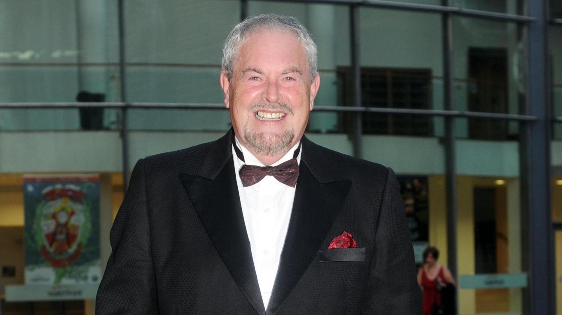 A man with short grey hair and facial hair wearing a tux with a brown bow tie.