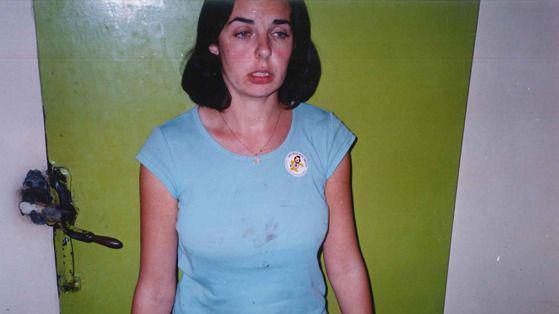 A woman in a stained blue t-shirt stands in front of a green painted wall looking shocked