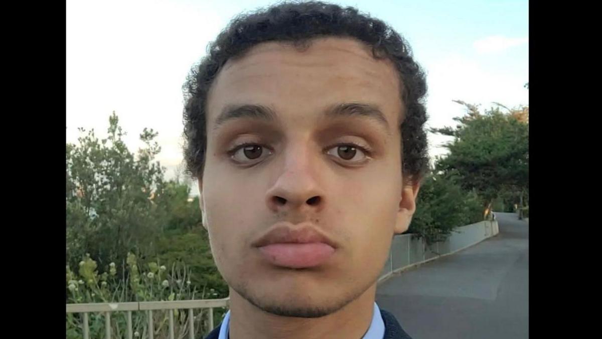 Adham Alkhatip with short curly black hair looks at the camera while recording an Instagram video. He is wearing a blue shirt and standing on a path with bushes in the background.