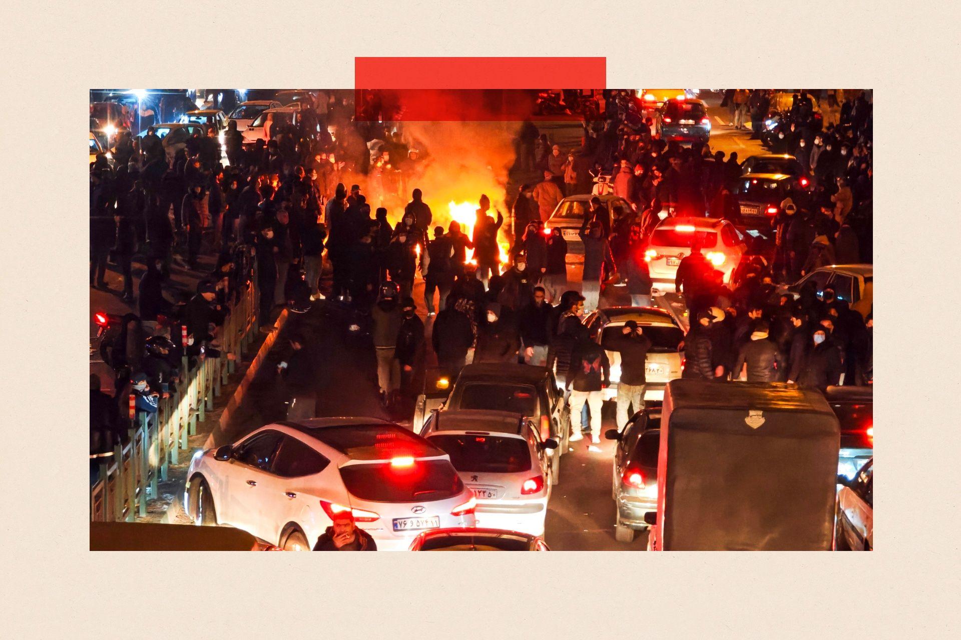 Hundreds of people out on the streets protesting, with a bonfire in the distance and cars in the foreground