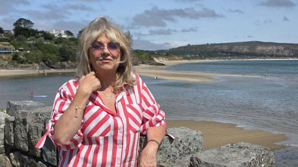 A blonde woman in a red and white striped shirt stands in front of a wall overlooking the seaside