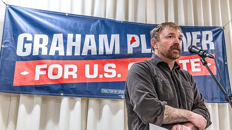 Graham Platner stands in front of a sign for his candidacy