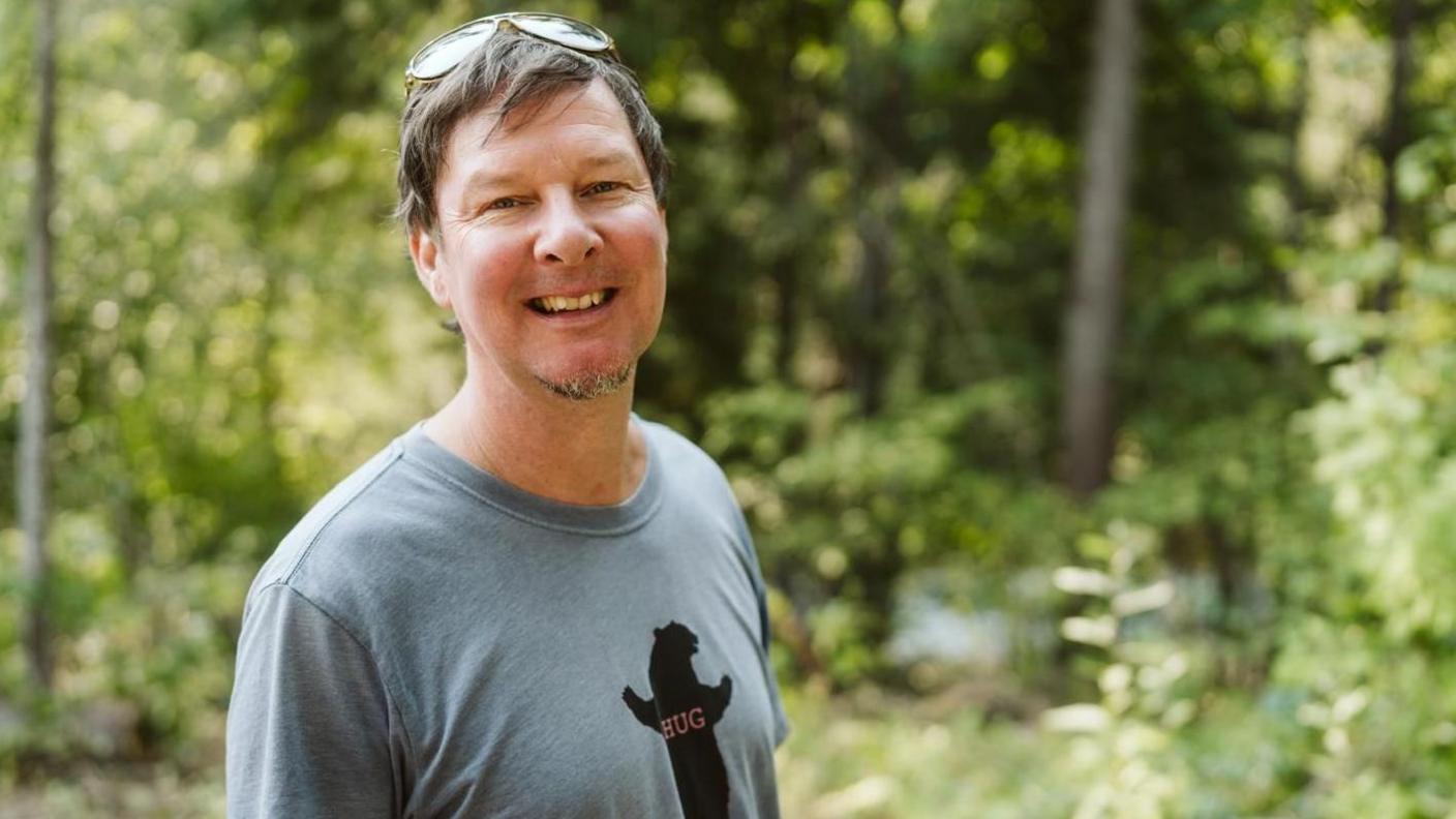 A mid shot of Lorne Warburton smiling while looking at the camera. He is wearing a grey shirt with a black bear bearing the words 'hug', with short brown and grey hair and sunglasses on his head. He is standing in front of an out of focus green wooded area.