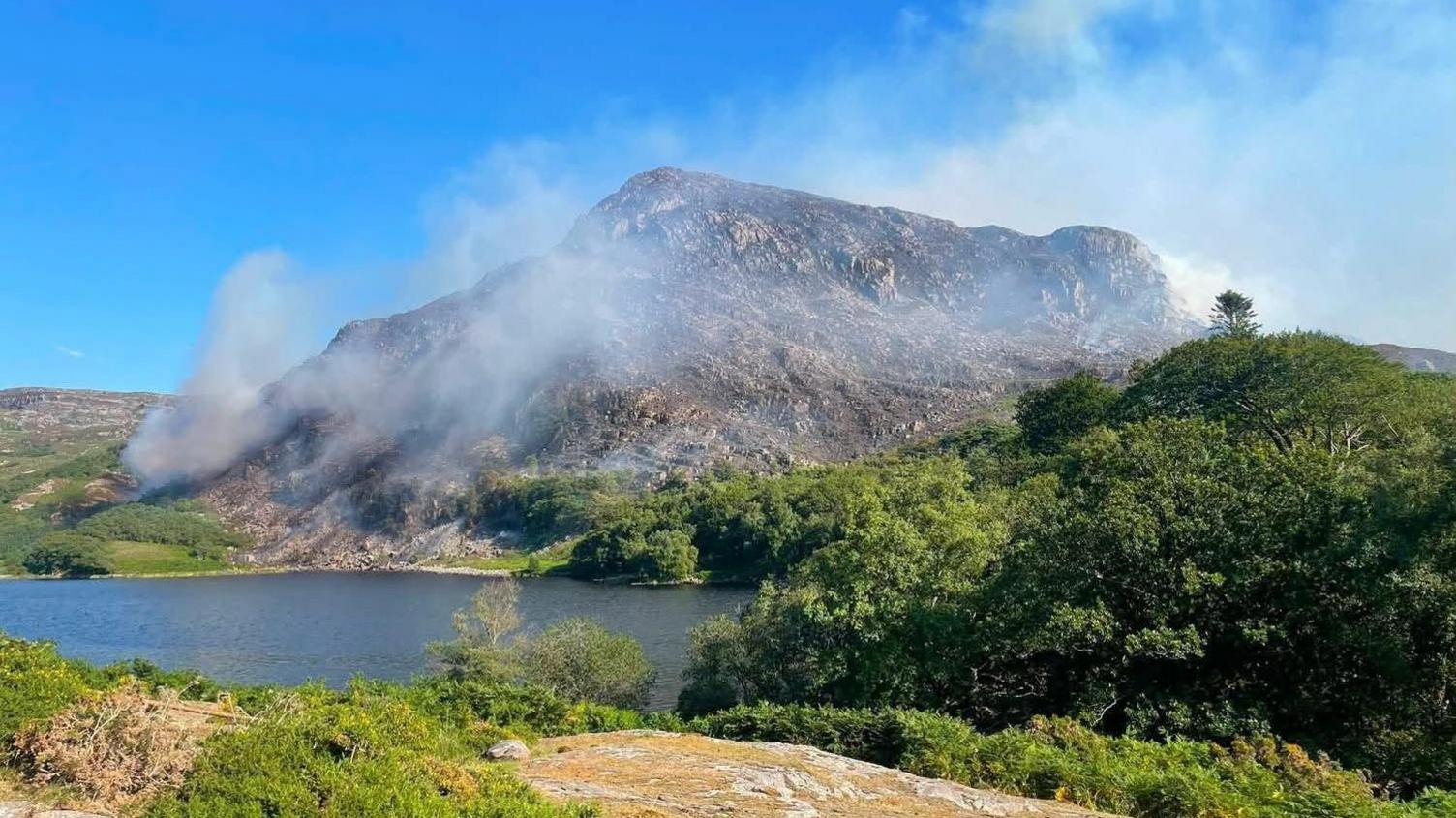 A scenic picture of a mountain with smoke rising from it. It is surrounding by grass and trees.