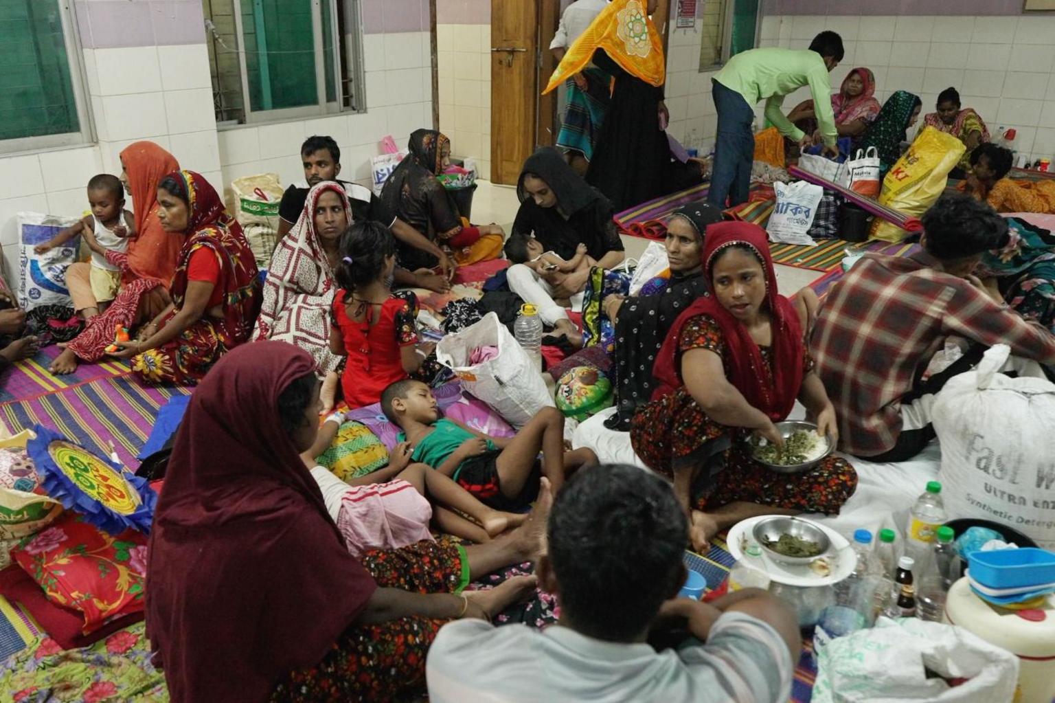 Overcrowded hospital ward in Mymensingh, Bangladesh, where dozens of people and children sit and lie on colourful mats on the floor, surrounded by bags and belongings, as the space appears stretched beyond capacity.