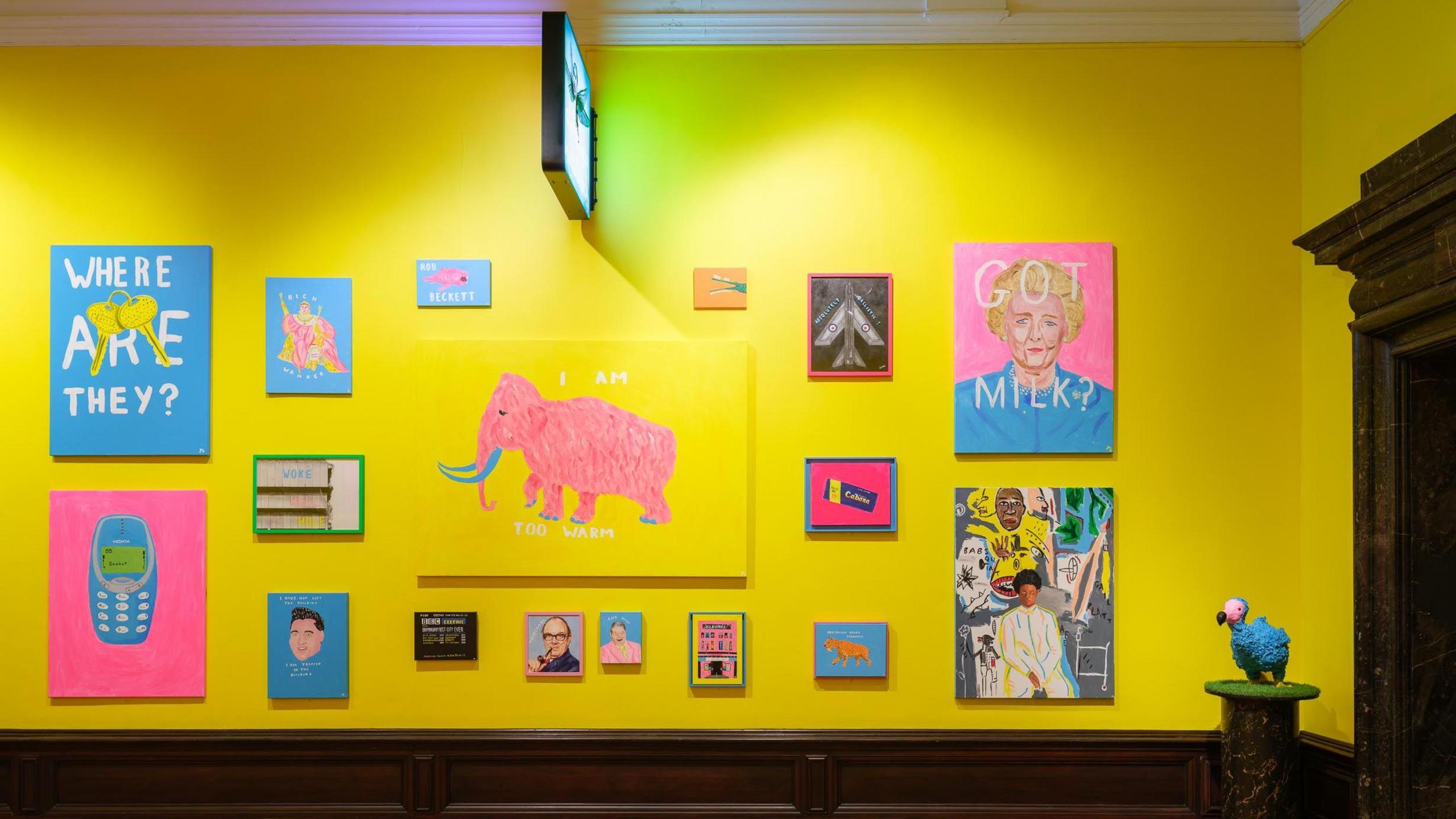 Colourful paintings largely in shades of blue and yellow hang on the yellow walls of an art gallery, with wooden floor. A sculpture of a dodo on a plinth lies to the right corner of the room.