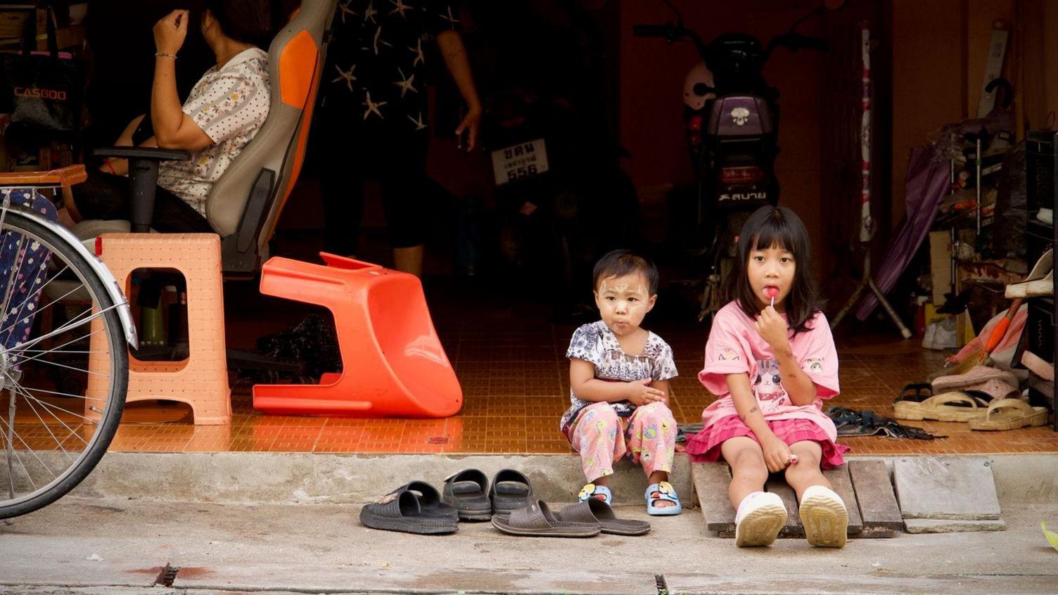 A young girl, lollipop in hand, sitting on the floor with another young child.