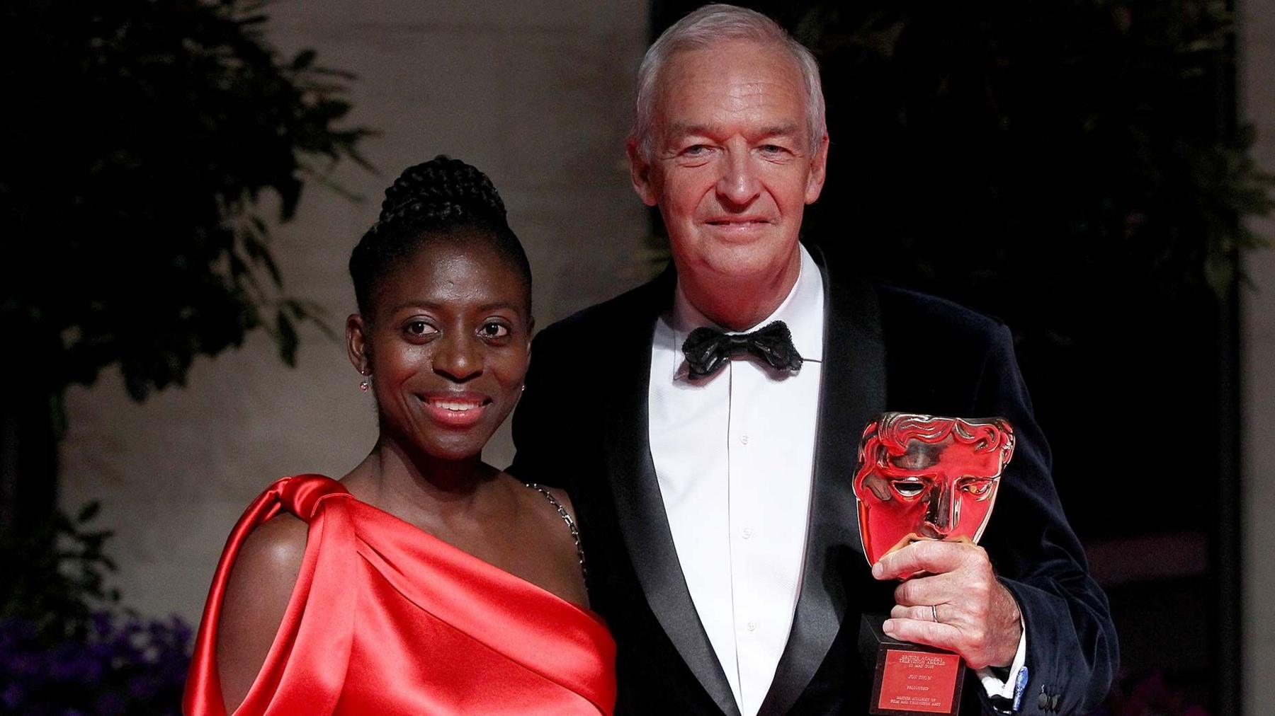 Jon Snow holding a Bafta and  standing with his wife Precious Lunga who wears a red red