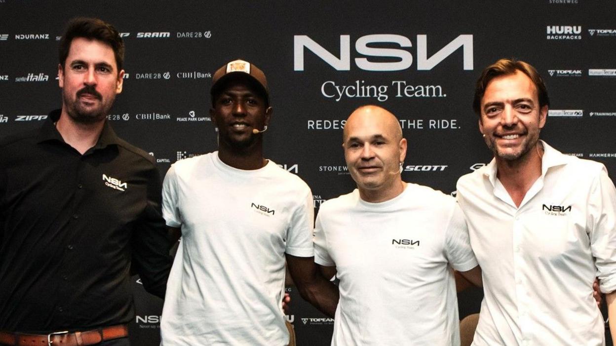 Biniam Girmay and Andres Iniesta wear white T-shirts as they stand in the centre of a line of four men, all with their arms around each other's waists. The man flanking Girmay on the left of the image wears a black shirt while the one on the right sports a white shirt. The four men, who are visible from the waist up, stand in front of a black board bearing the slogan NSN Cycling Team and other sponsor logos, while all four also have NSN logos on their left breasts