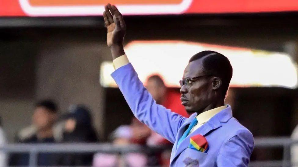 Smartly-dressed Michel Kuka Mboladinga, better known as Lumumba Vea or the "living statue", stands still with his armed raised while watching his country DR Congo