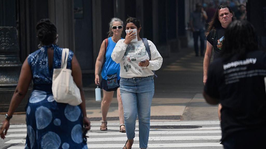 A woman in a white sweatshirt and jeans holds a cloth to her face as she crosses a city street, alongside other pedestrians.