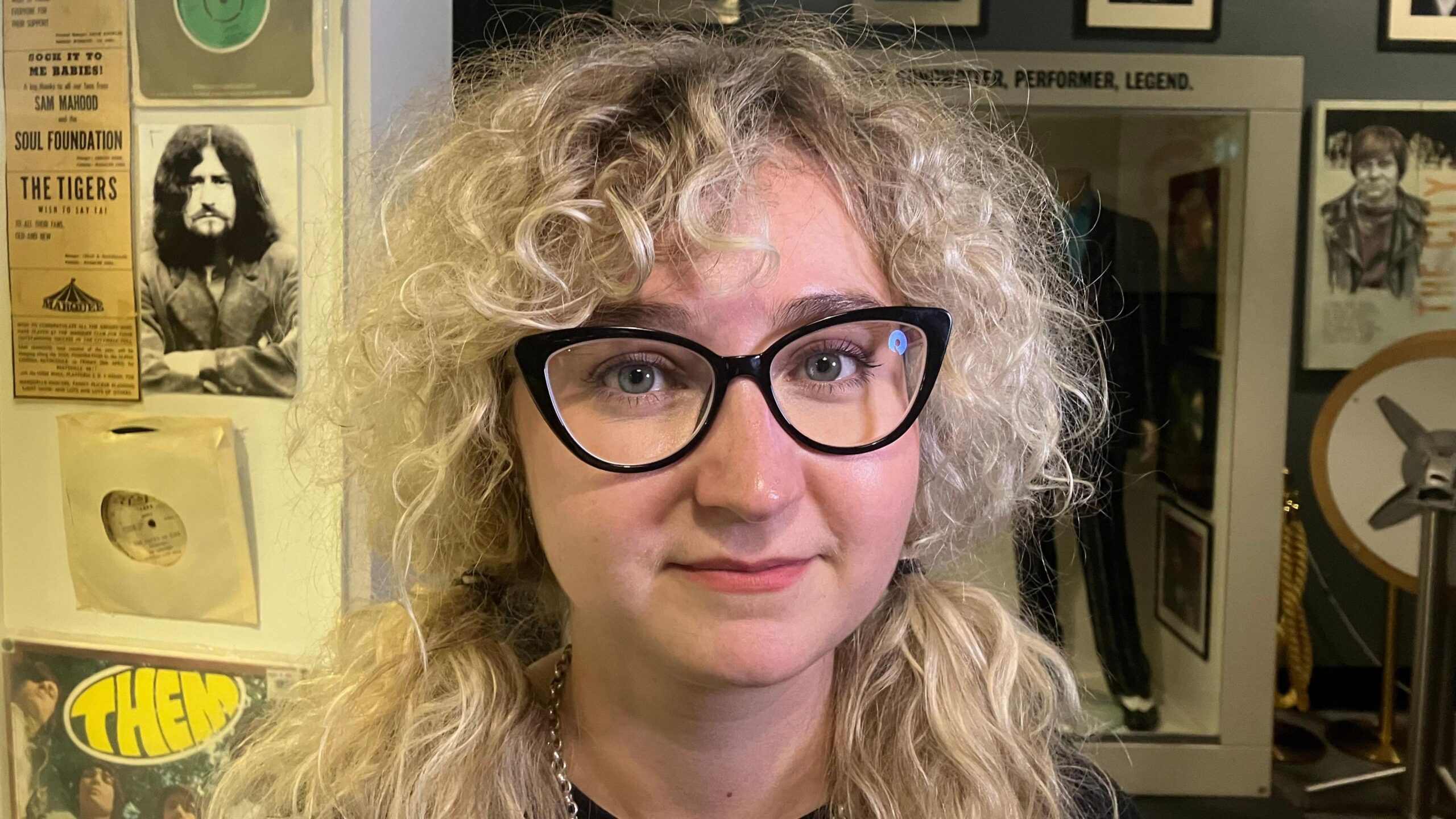 A woman with long, blonde, curly hair. She is wearing black glasses.