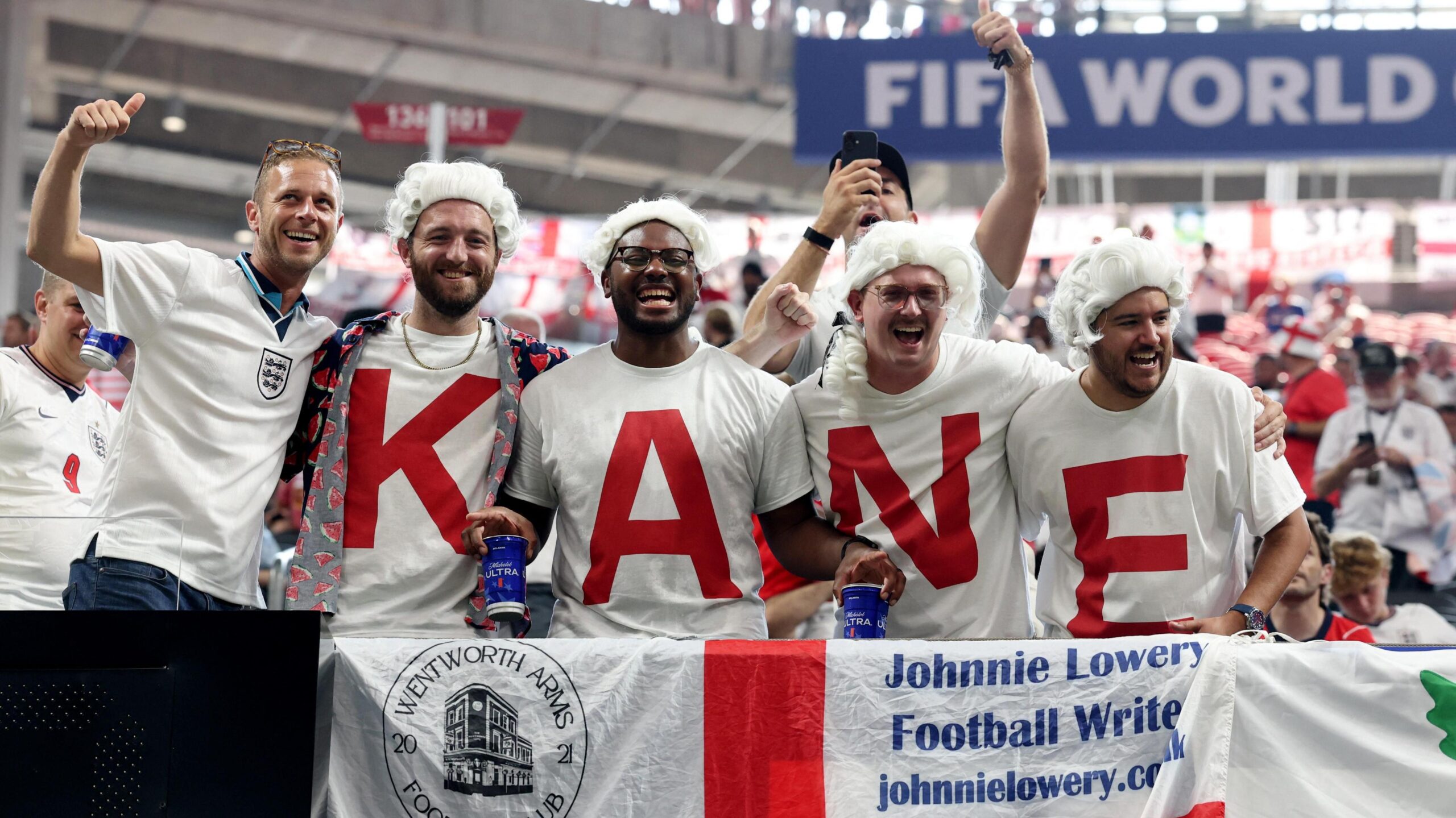 England fans display the name of Harry Kane on their shirts inside a stadium. They are all wearing white wigs.