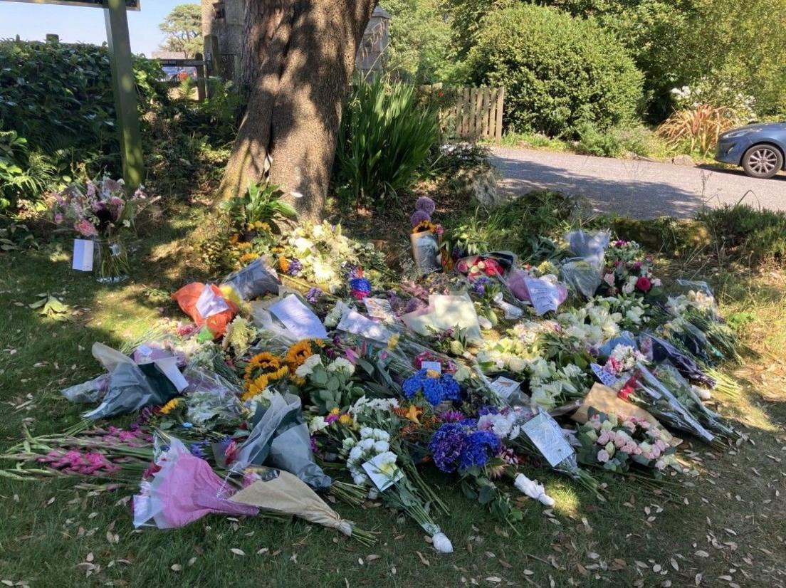 Dozens of bouquets of flowers laid on the grass in front of a tree near a hotel. Among the floral tributes at the back is a black and white photo of Widdecombe.
